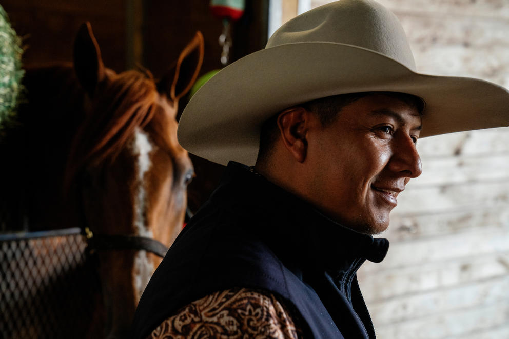 A close up portrait of a man wearing a cowboy hat. A horse is in a stable behind him.