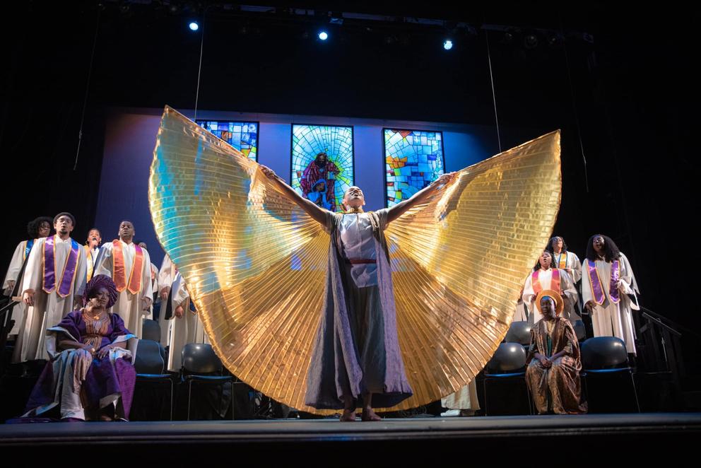 An angel dancer with yellow fabric wings performs onstage in front of a gospel choir