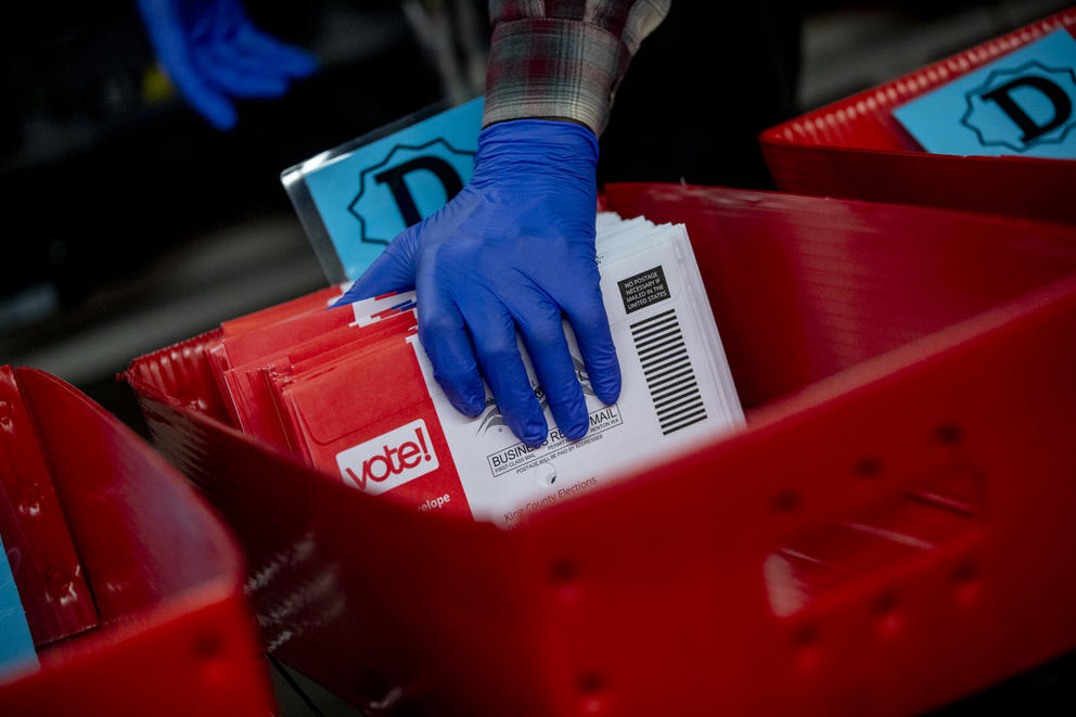 A hand holds election ballots in their mailing envelops in a bin.