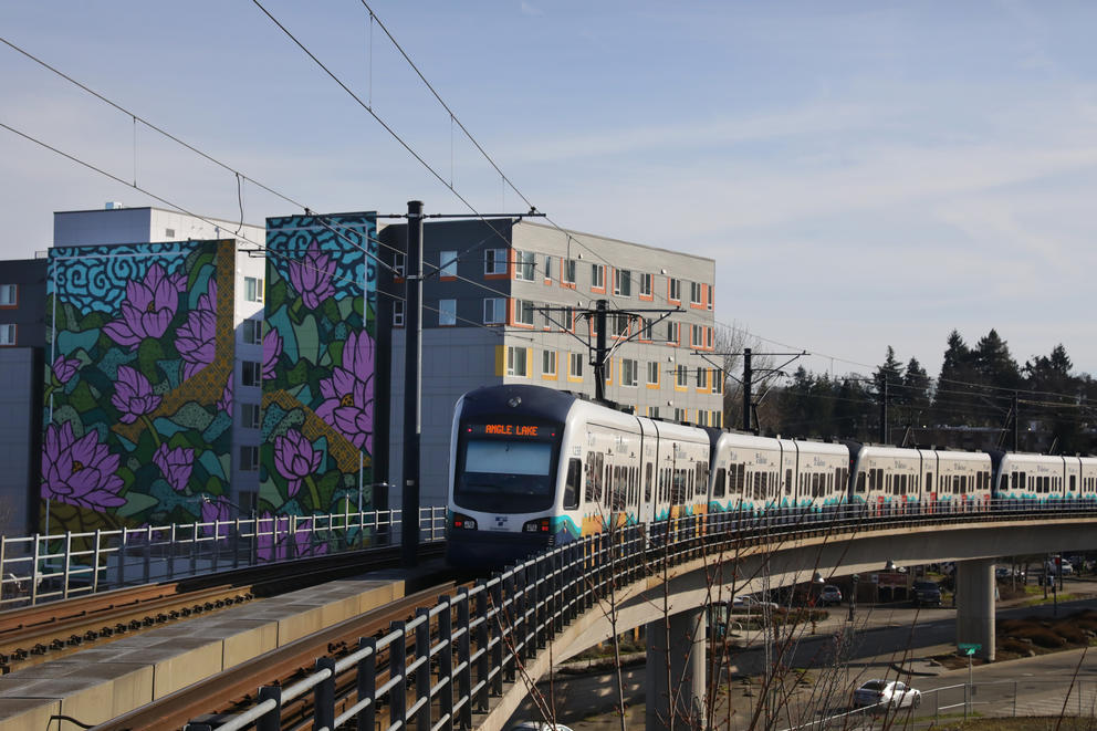 a light rail train pulls into a station on elevated tracks. it's passing an apartmnet building with a colorful flower mural painted on the side