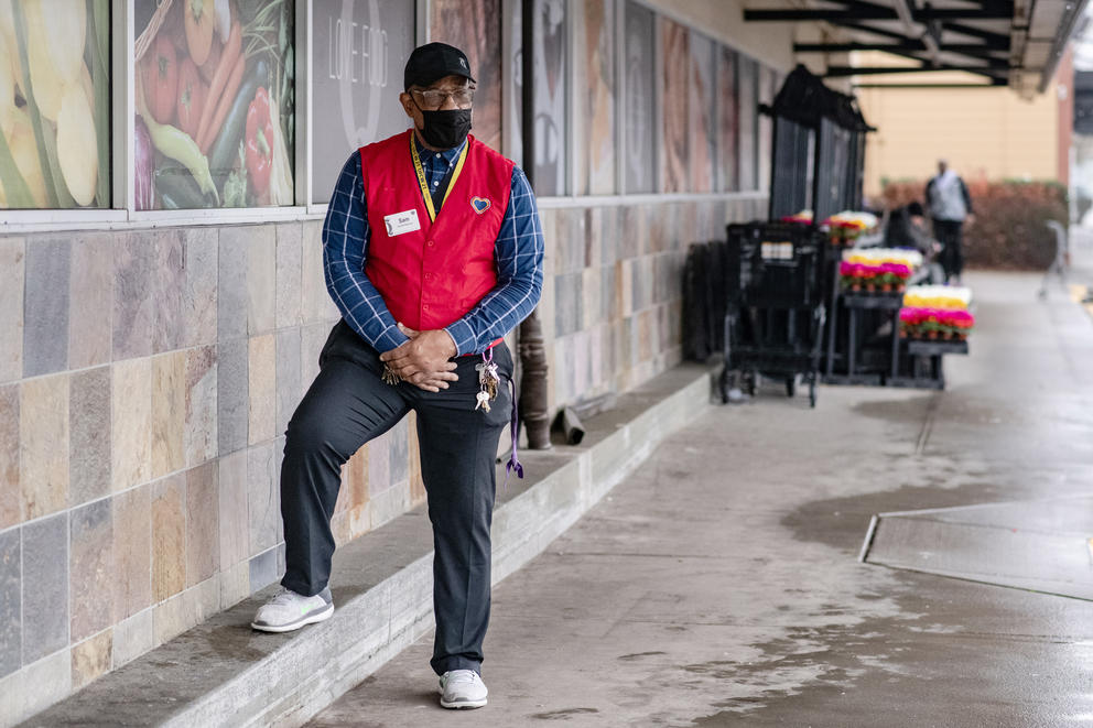 A grocery store manager in a red vest poses for a portrait outside the store.