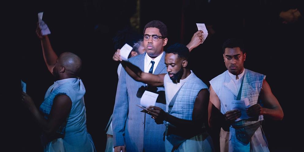 a scene from a play featuring Black men in suits waving small pieces of paper
