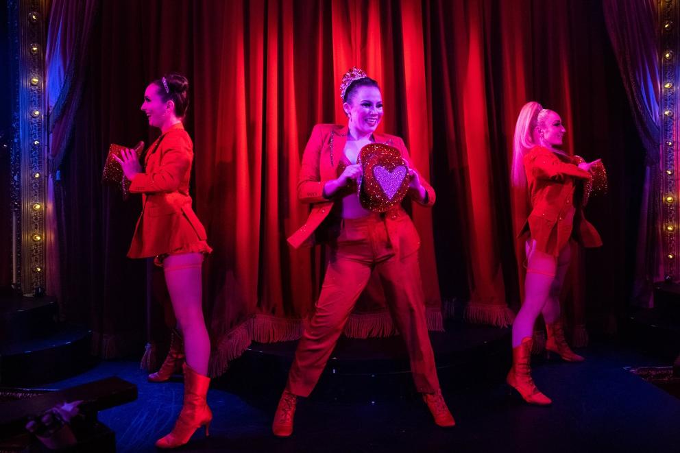 three cabaret performers in red suits on stage in deep red lighting