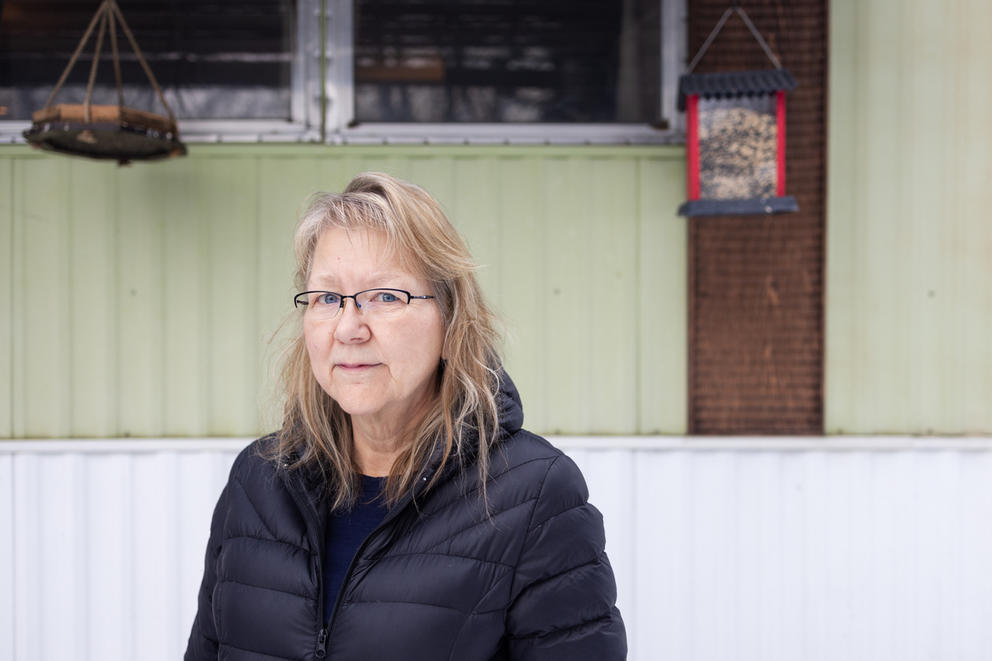 A woman poses for a photo in front of her home at Sun Tides mobile home community in Gleed, Washington. She's wearing a black puffer jacket and black half rim glasses.