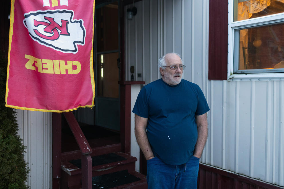 A man stands outside his home in Moxie Community. He's wearing a black short sleeve shirt, wire frame glasses and jeans. Next to him is a Kansas City Chiefs flag.