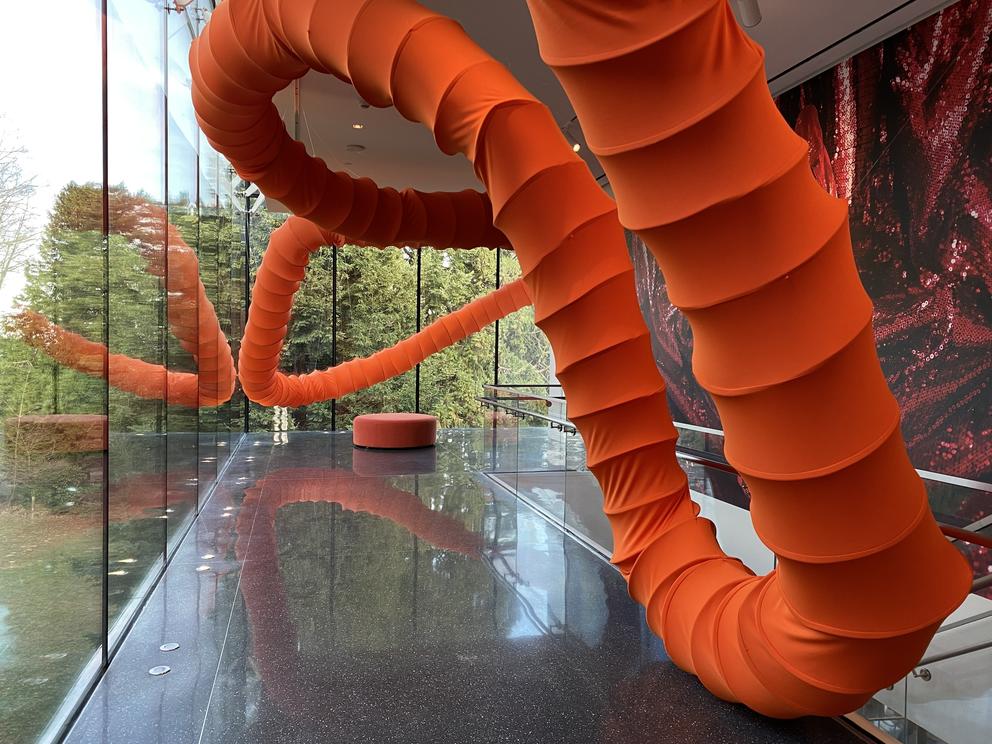 photo of a museum installation of a long orange ringed tube stretching across a windowed gallery