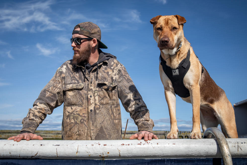 Green in a camo jacket stands at a rail next to his light brown dog