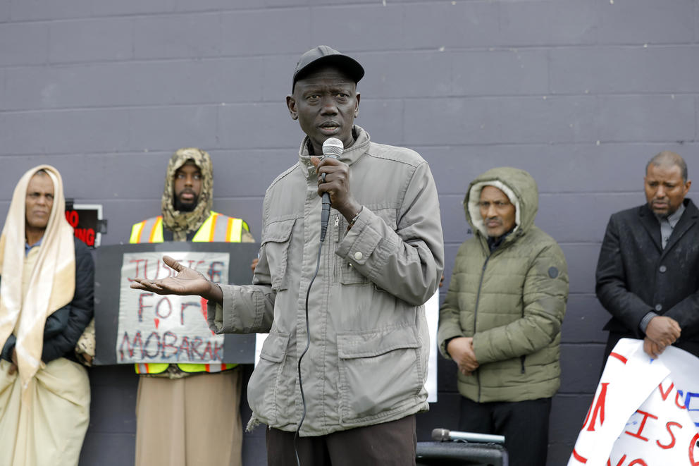 a man speaks into a microphone at a rally with people holding signs in the bacground