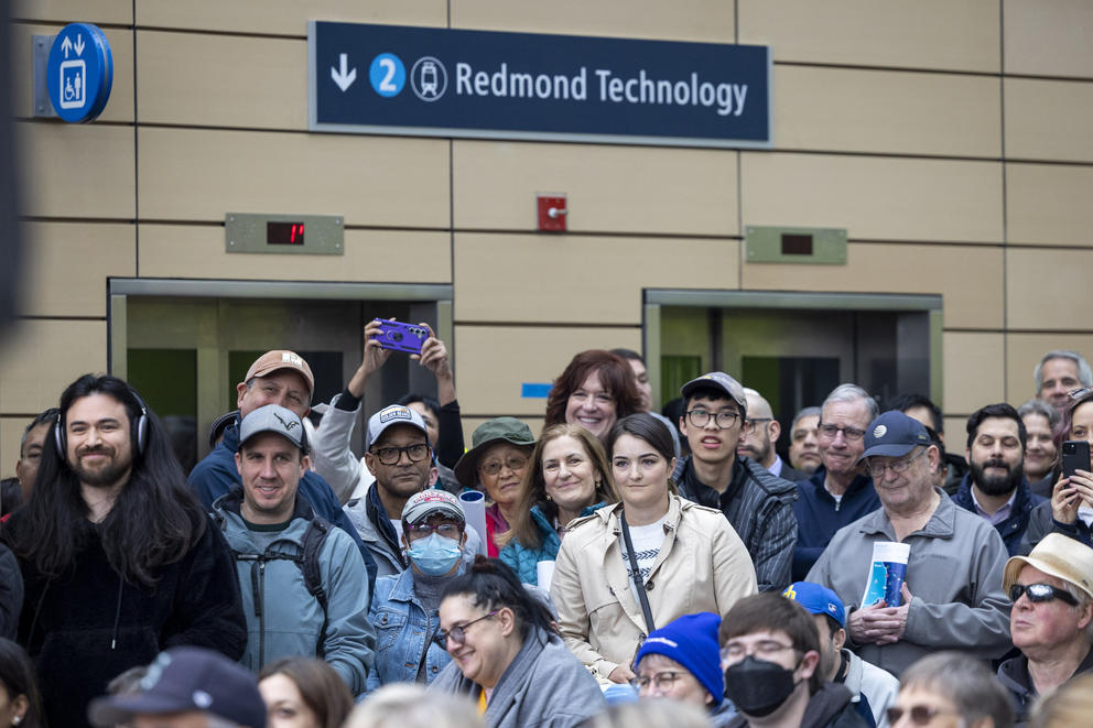 An attentive crowd is listening to someone off camera. In the background there is a sign that says "2 Redmond Technology," indicating the direction of the train.