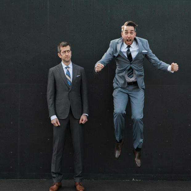 A white man with short, dark hair wearing a blue suit and light brown shoes is mid-jump with an excited expression on his face. Another man in a darker suit and dark brown shoes stand next to him, looking up at him quizzically. There is a dark grey wall behind them.. 