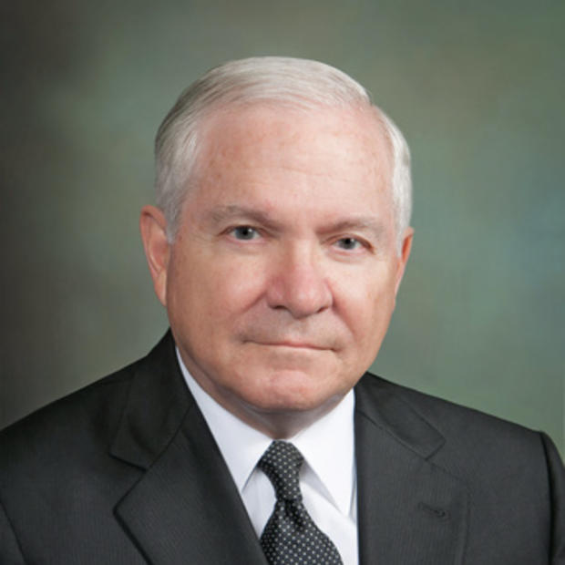 White man with mostly white short hair wearing a white button-down, dark tie and suit jacket against a grey background