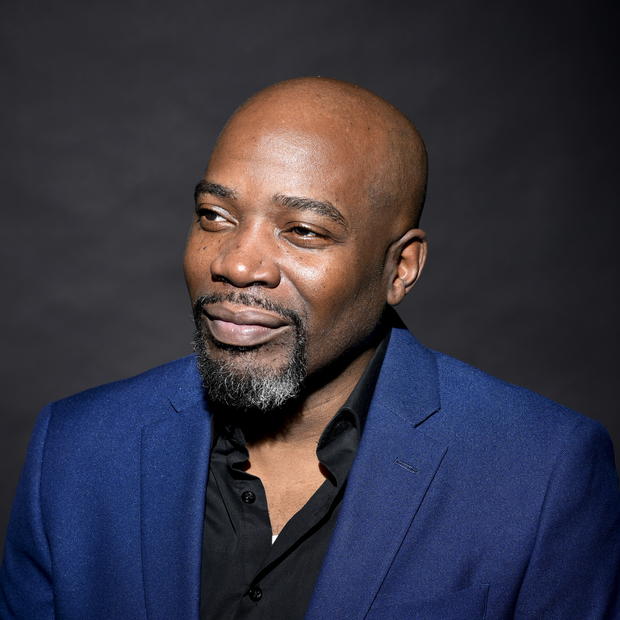 Black man with goatee wearing a black button-down shirt and royal blue blazer looks off to the side against a dark grey background