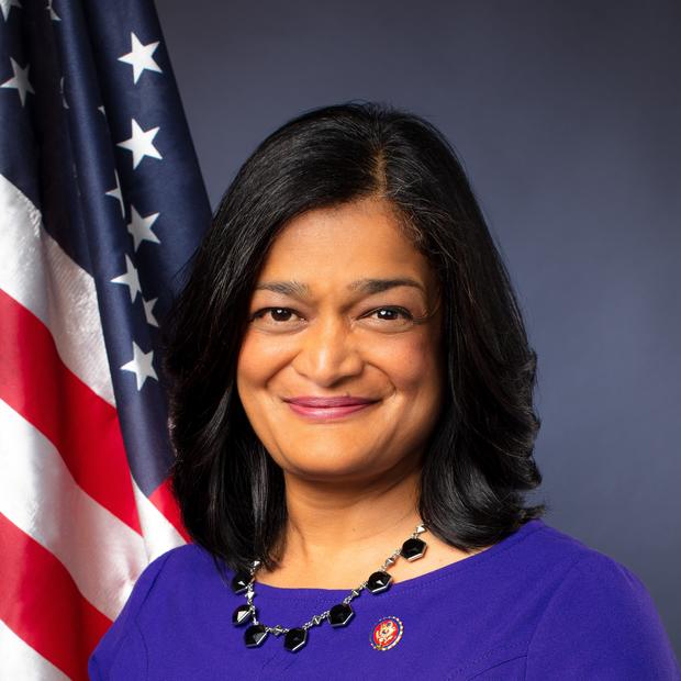 Woman with light brown skin and dark, shoulder-length straight hair wearing a royal blue top and beaded necklace with an American flag and grey background