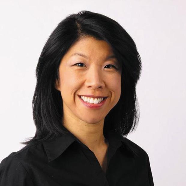 Asian woman with dark, shoulder-length hair wearing a black button-down top smiles at the camera against a white background