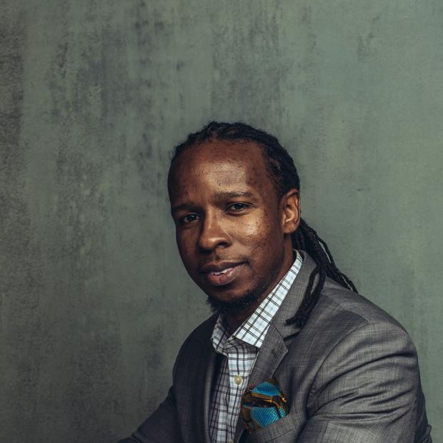Black man with long, thin dreadlocks pulled back into a low ponytail, wearing a checked button-down shirt and grey blazer shits at a table covered with a cream tablecloth against a grey background