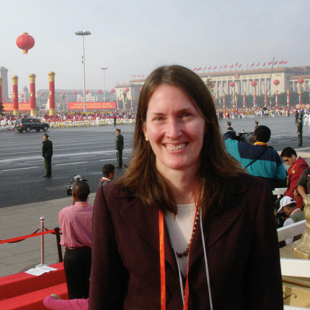 White woman with straight, shoulder-length brown hair wearing a dark blazer over a white shirt with beaded necklace and orange lanyard stands smiling at the camera. There is an outdoor, formal government celebration in another country taking place in the background