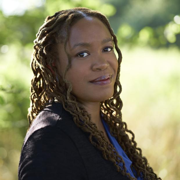 Black woman with thin, curled, dreadlocks pulled halfway up stands sideways in a field, looking over her shoulder at the camera. She is wearing a blue shirt and dark blazer. 