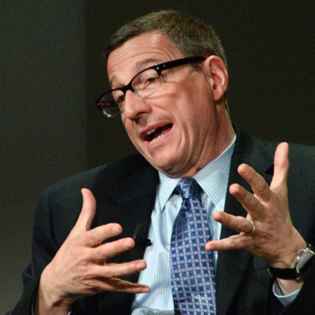 White man with short, dark hair wearing glasses, a light blue button-down, dark blue necktie and dark blazer is gesticulating with his hands in front of his body as he is speaking