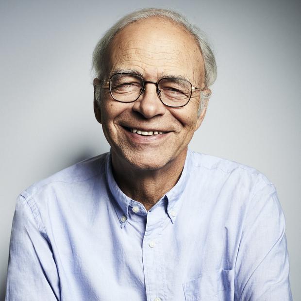 Older white man with short, white hair and glasses wearing a light blue button-down stands with his arms crossed and smiles at the camera against a light grey backdrop