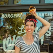 A white woman with dark, curly hair pulled back into a bun on the top of her head  holds a sandwich above her head. She is wearing a black and white striped t-shirt and red bandana as a headband.  She is smiling and looking up at the sandwich. In the background is a restaurant's glass window advertising burgers and milkshakes. 