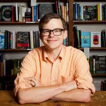 White man with short dark hair wearing glasses and a peach-colored button-down top has his arms crossed and resting on a table. He is smiling at the camera and there are bookshelves filled with books in the background