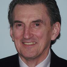 Older white man with combed back dark grey hair wearing a white striped button down, dark necktie and dark suit jacket smiles at the camera against a light grey backdrop