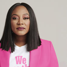 Black woman with dark, shoulder-length straight hair wearing a gold nose ring, a white t-shirt with the word "We" visible, and pink blazer against a light grey background