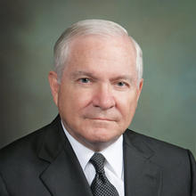 White man with mostly white short hair wearing a white button-down, dark tie and suit jacket against a grey background
