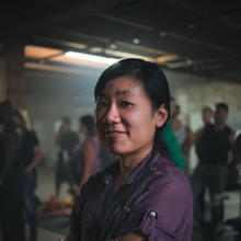 Asian woman with dark hair pulled back wearing a deep purple short-sleeve top stand sideways with her arms crossed. She is in a dark room with several people standing in the background