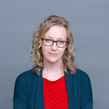 White woman with shoulder length wavy hear wearing glasses, a red shirt with a geometric design and navy blue cardigan against light grey background