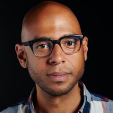 Black man with slight growth of facial hair wearing dark glasses and a blue and white checkered button-down shirt against a black background