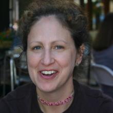 White woman with brown curly hair pulled into an updo wearing a beaded necklace and dark shirt smiles at the camera. She is sitting in a restaurant. 