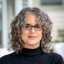 White woman with salt and pepper shoulder-length, curly hair wearing glasses and a black turtleneck with a blurred background