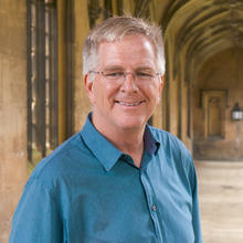 White man with short, grey hair wearing glasses and a blue button-down shirt smiles at the camera. He is standing underneath the stone arches of a long hallway of an historic building
