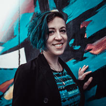 White woman with dark hair in a bob style wearing a striped blue and black shirt under a black blazer standing in front of a wall with broad brush-type strokes of blue, black and brownish purple