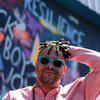 Black man with short dreadlocks and a goatee wears blue sunglasses and a pink button-down shirt. He is standing with his left hand on the back of his head. In the background there is a wall with colorful graffiti. 