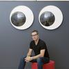 White man with dark short hair wearing glasses, a wristwatch, a black t-shirt and jeans sits on a round red cushion with a blue wall and a large pair of googly eyes in the background