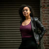 Black woman with dark, shoulder-length curly hair and glasses wearing a purple top and black leather jacket stands with her hands on her hips against a brick wall with an industrial style garage door