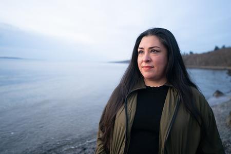 Person in front of body of water. She is wearing black clothing, has long dark hair and wears a greenish scarf