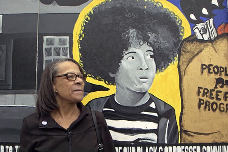 A woman stands in front of a Black Panther Party themed mural, featuring a person with afro, in Seattle.