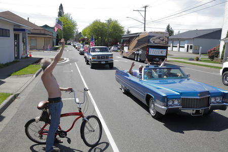 A man on a bicycle waves an American flag at a car driving by in a parade