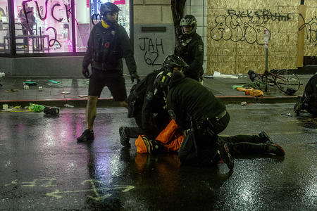 A police officer kneels on a detained person's neck on wet pavement on a dark street in Downtown Seattle