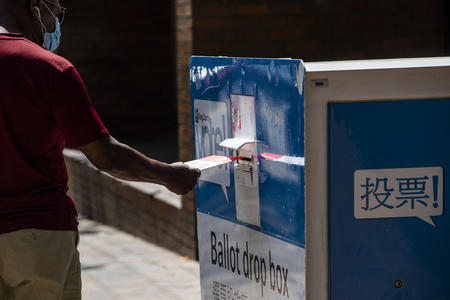 A person in a mask puts a ballot in a ballot box, with back to camera