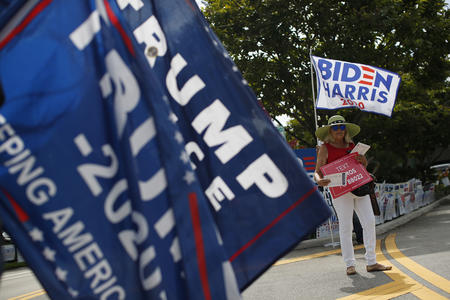 Woman holding a Joe Biden sign is partially obscured by a Trump 2020 flag.