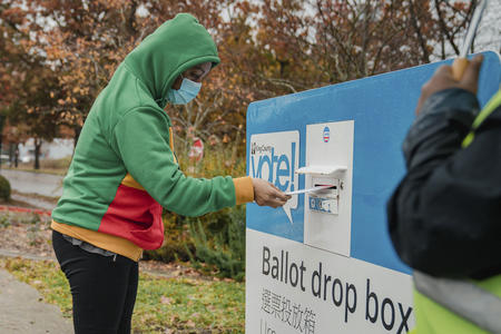 Person in green hoodie puts a ballot in a blue and white ballot drop box