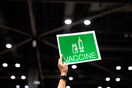 A hand holds up a sign with a graphic of a syringe and bottle with the word "Vaccine" beneath it