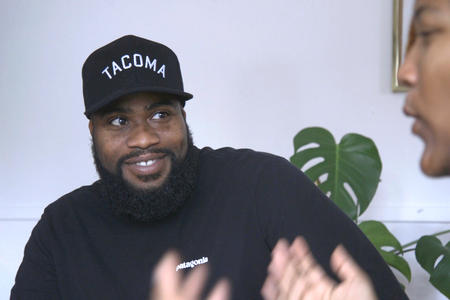 A man in a black “Tacoma” hat smiles as a woman talks with him