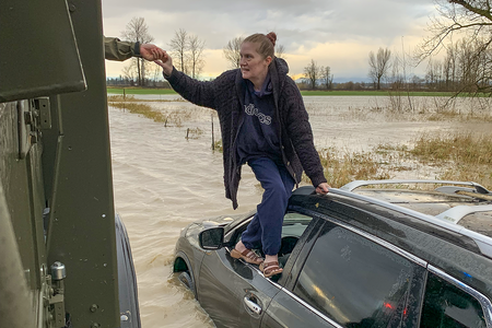 A woman balances on a car door's window well and grasps a hand reaching for her. The car is mostly submerged in floodwater