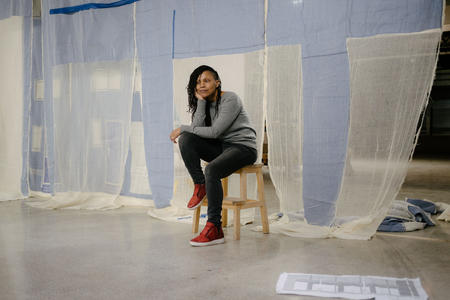 Person on a small ladder looking at the camera, in front of white and blue sheets of fabric that are semi-transparent