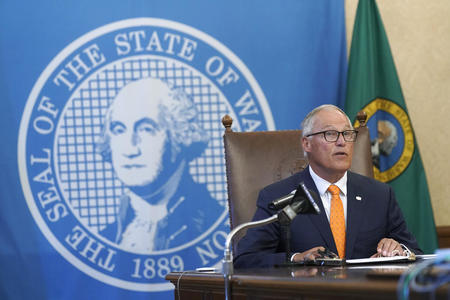 Gov. Inslee sits in front of a WA state seal banner at a press conference